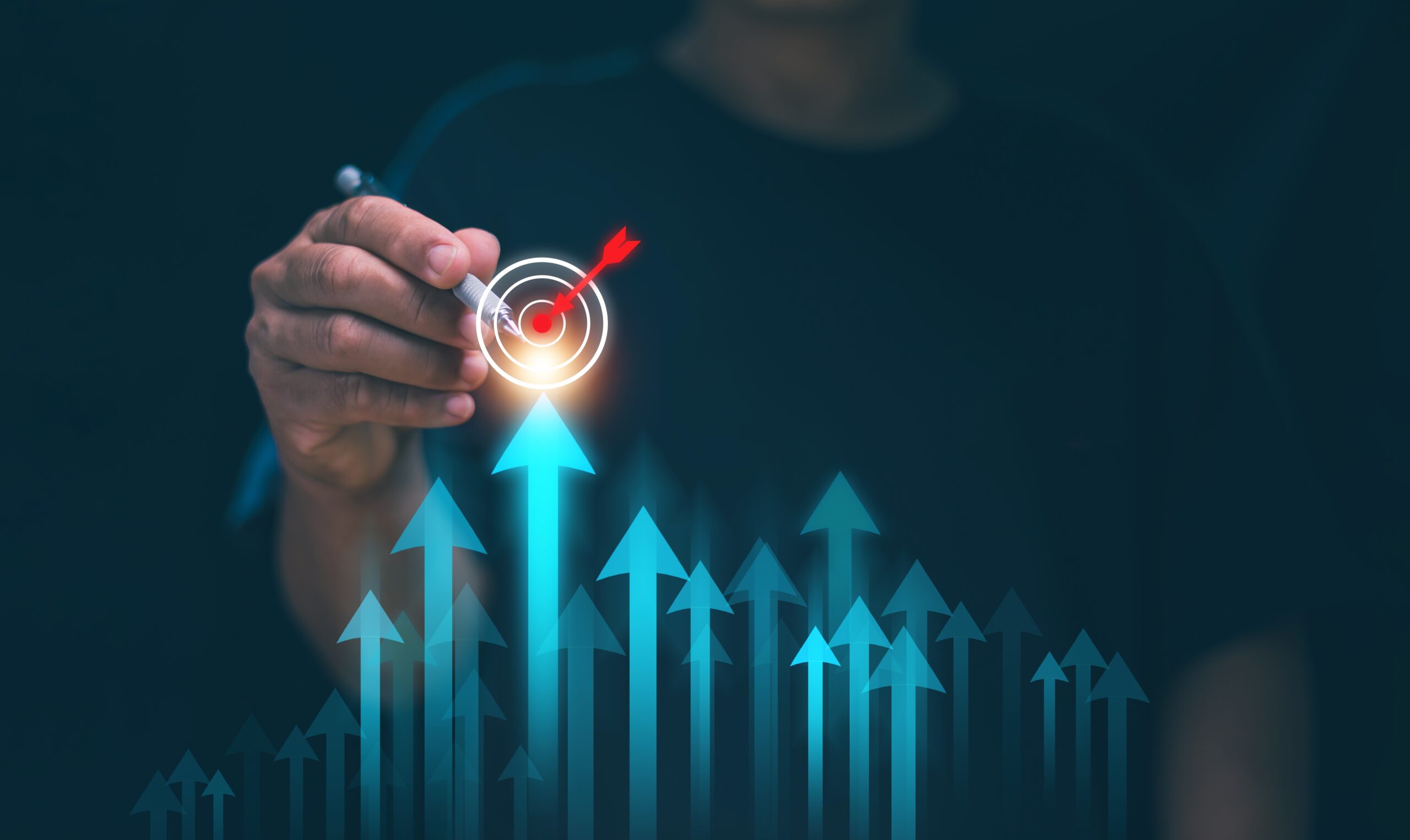 A person in a dark shirt holds a glowing target with a red arrow at its center above a cluster of upward-pointing, illuminated blue arrows of varying heights. The scene is set against a dark background, emphasizing the concepts of achieving goals and business growth.